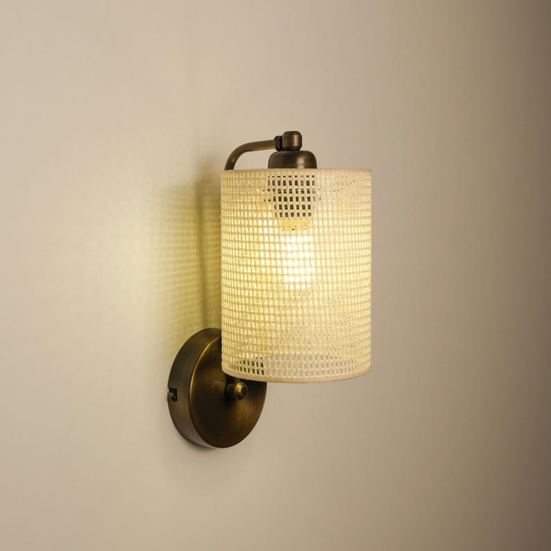 May include: A wall-mounted light fixture with a cylindrical, off-white shade featuring a grid pattern. The fixture has a bronze-colored base and arm, and the light is illuminated. The design is simple and modern.