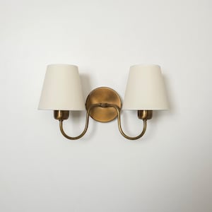 May include: A double sconce light fixture with a brass finish. It features two off-white, cone-shaped lampshades. The fixture is mounted on a white wall, showcasing a classic design.