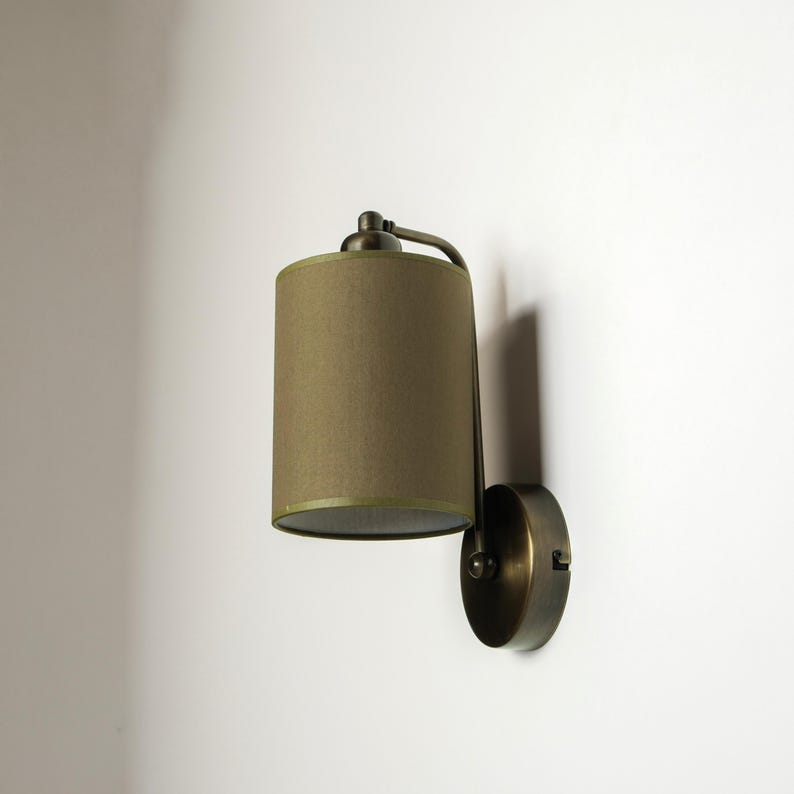 May include: A wall-mounted lamp with a cylindrical olive green lampshade. The lamp has a curved arm and a circular base, all in a bronze finish. The lamp is designed to be mounted on a wall.