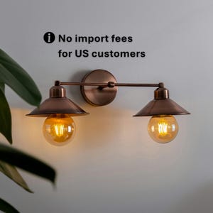 Könnte beinhalten: Eine bronzefarbene Wandleuchte mit zwei Lichtern im Vintage-Industriedesign. Jedes Licht hat einen konischen Schirm und eine runde, freiliegende Glühbirne. Der Text "No import fees for US customers" ist sichtbar.