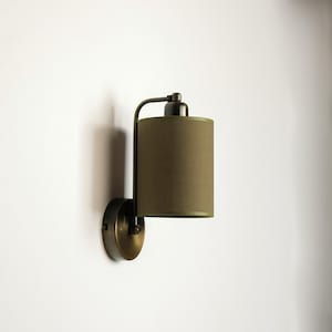 May include: A wall-mounted lamp with a cylindrical olive green lampshade. The lamp has a dark metal finish and a curved arm. The shade is a solid color, and the lamp is mounted on a round base. The lamp is designed for indoor use.