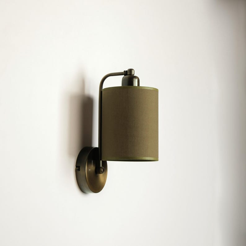 May include: A wall-mounted lamp with a cylindrical olive green lampshade. The lamp has a dark metal finish and a curved arm. The shade is a solid color, and the lamp is mounted on a round base. The lamp is designed for indoor use.