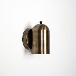 Peut inclure: Applique murale en laiton antique. Le luminaire a un abat-jour arrondi en forme de cloche avec des lignes verticales et un petit bouton décoratif. La base circulaire est fixée à une plaque murale.