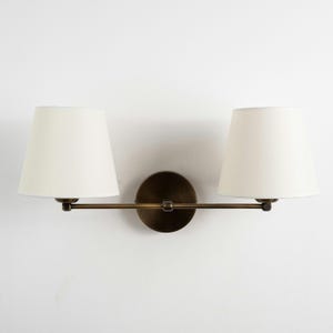 Op de afbeelding: Een dubbele wandlamp met een bronzen afwerking. Het heeft twee off-white, kegelvormige lampenkappen. De lamp heeft een cirkelvormige achterplaat en een horizontale balk om de kappen vast te houden. Een klassieke lamp.