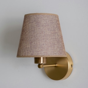 May include: A wall-mounted sconce with a conical lampshade. The lampshade is made of a textured, light brown fabric with a darker brown trim. The fixture has a brass finish, including the round base and the arm holding the shade.