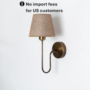 May include: A wall-mounted lamp with a tan fabric shade and a bronze-colored metal frame. The lamp has a curved arm and a circular base. The text in the image reads: No import fees for US customers.