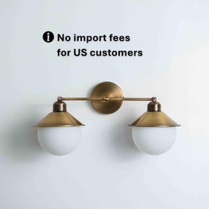 May include: A brass-toned wall sconce with two lights. Each light has a white globe and a conical, gold-colored shade. The fixture is mounted on a round backplate and has a horizontal bar connecting the lights. Text: No import fees for US customers.