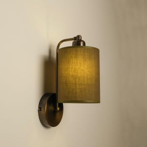 May include: A wall-mounted lamp with a cylindrical olive green lampshade. The lamp has a bronze-colored metal frame and a circular base. The shade is made of a textured fabric, and the lamp is casting a warm glow.