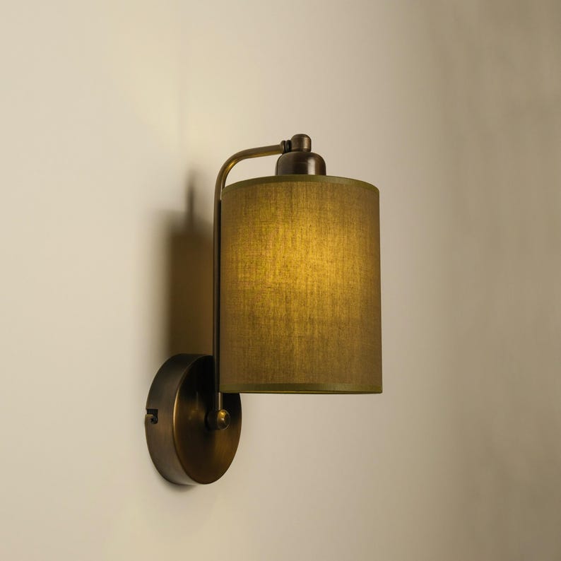 May include: A wall-mounted lamp with a cylindrical olive green lampshade. The lamp has a bronze-colored metal frame and a circular base. The shade is made of a textured fabric, and the lamp is casting a warm glow.