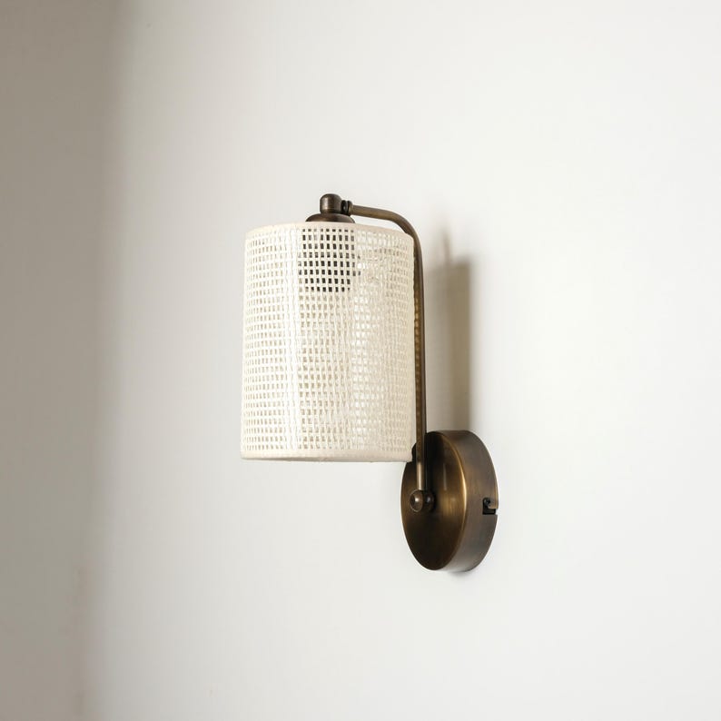 May include: A wall-mounted light fixture with a cylindrical, woven white shade. The fixture has a dark bronze finish and a curved arm connecting the shade to a circular base. The design is minimalist and suitable for interior lighting.