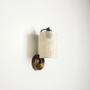 May include: A wall-mounted light fixture with a cylindrical, off-white woven shade. The fixture has a dark bronze finish on the base and supporting arm. The shade is a textured, open weave design.