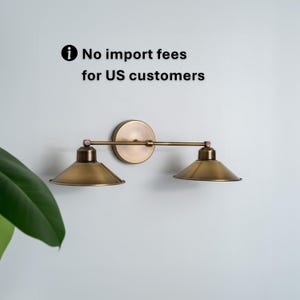 May include: A brass-toned wall sconce with two conical shades. The fixture has a circular backplate and a horizontal arm connecting the shades. The text "No import fees for US customers" is visible.