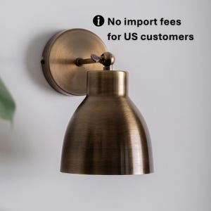 Op de afbeelding: Een antiek messing wandlamp met een koepelvormige kap. De lamp heeft een cirkelvormige achterplaat en een draaibare arm. De tekst in de afbeelding luidt: Geen importkosten voor Amerikaanse klanten.