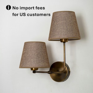 May include: A wall-mounted double lamp fixture with two brown fabric shades. The fixture has a brass finish and a circular base. The text "No import fees for US customers" is visible.