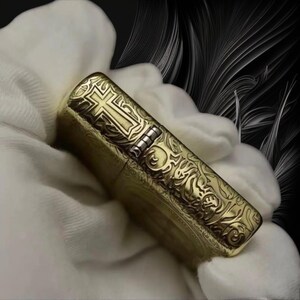 May include: A gold-colored metal lighter with an engraved cross design on the front and ornate patterns on the sides. The lighter has a silver hinge and is held against a white fabric background.