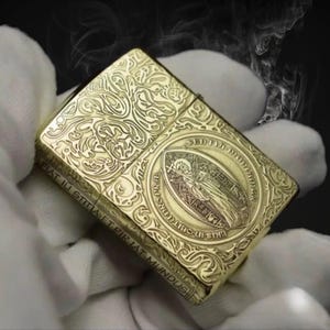 May include: A gold-colored, rectangular lighter with intricate engraved patterns. The design includes floral motifs and a central oval emblem with a religious figure and text. The lighter is held against a dark background with wisps of smoke.