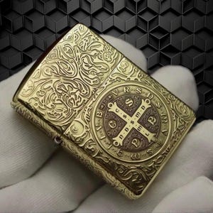 May include: A gold-colored, rectangular lighter with intricate engraved patterns. The lighter features a detailed cross design within a circular frame on the front, surrounded by text. The overall design is ornate and vintage-inspired.