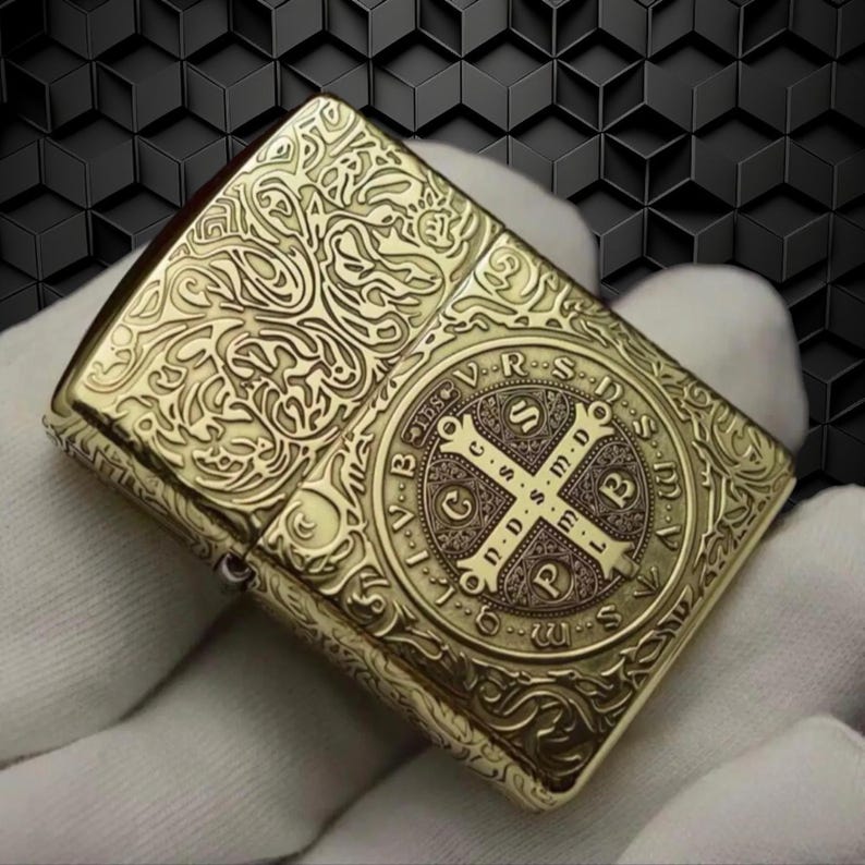 May include: A gold-colored, rectangular lighter with intricate engraved patterns. The lighter features a detailed cross design within a circular frame on the front, surrounded by text. The overall design is ornate and vintage-inspired.