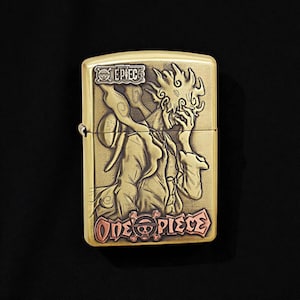 May include: A brass-coloured lighter with an engraved illustration of a character from the anime series "One Piece." The design includes the text "EPIC" and "ONE PIECE" in a stylized font. The lighter has a classic rectangular shape.