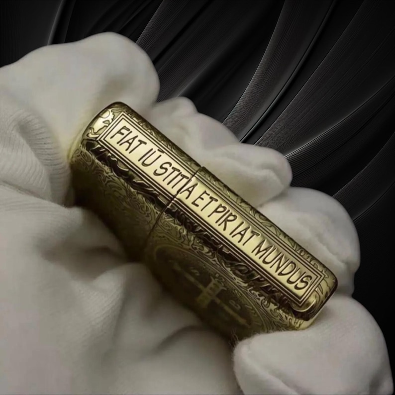 May include: A gold-colored lighter with intricate engraved patterns and the inscription "FIAT IU STITIA ET PIR IAT MUNDUS". The lighter is held in a white glove, with a dark background.