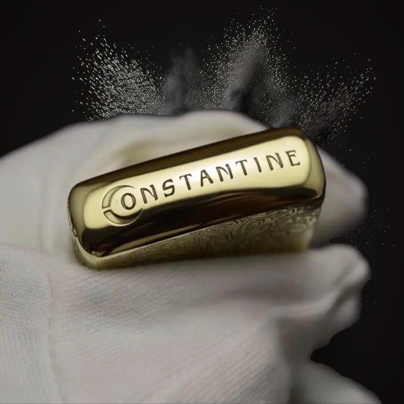 May include: A gold-colored rectangular lighter with the word "CONSTANTINE" engraved on it. The lighter has a decorative pattern on its side and is set against a dark background.