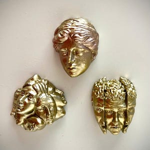 Puede incluir: Tres esculturas de pared de color dorado con rostros humanos. La pieza superior muestra un solo rostro con cabello ondulado. La pieza inferior izquierda muestra un grupo de varios rostros. La pieza inferior derecha muestra un rostro segmentado.