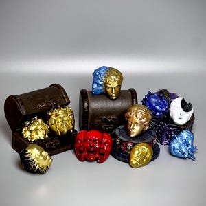 Puede incluir: Una colección de pequeñas figuras esculpidas de rostros humanos en varios colores, incluyendo oro, rojo, azul y blanco. Algunos rostros están agrupados, mientras que otros se colocan sobre o dentro de pequeños cofres de madera oscuros.