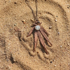 May include: A copper-colored pendant necklace with a central, round, light-colored stone. The pendant has a starburst design with multiple pointed elements. The necklace rests on a sandy surface.