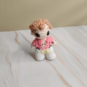 Op de afbeelding: Een handgemaakte gehaakte pop met lichtbruin haar, zwarte ogen en een roze shirt. De pop draagt een witte broek met gele accenten en zwarte bretels. De pop staat rechtop.
