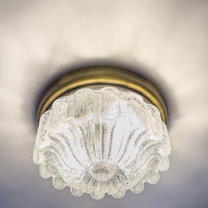 Pode incluir: Uma luminária de teto de vidro transparente texturizado com uma base de metal cor de latão. A luz tem um design em forma de flor com uma borda ondulada. A luminária é montada em um teto branco.
