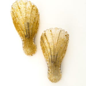 May include: A pair of wall sconces shaped like seashells, crafted from clear glass with embedded gold-coloured flecks. Each fixture features a ribbed, fan-like texture radiating from a central circular mounting point, creating a translucent appearance.