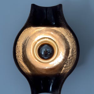 Könnte beinhalten: Dunkelbraune Wandleuchte mit strukturierter, goldfarbener Innenseite. Die Leuchte hat eine markante, geschwungene Form mit einer zentralen Lichtquelle. Das Licht reflektiert sich im goldenen Inneren und erzeugt einen warmen Schein.