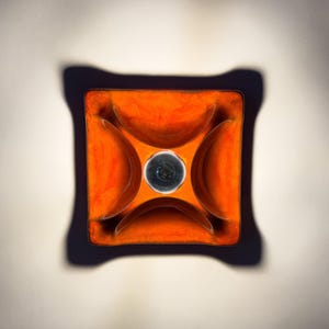 Könnte beinhalten: Ein orangefarbener quadratischer Aschenbecher mit dunklem Rand und vier gebogenen Vertiefungen. In der Mitte befindet sich eine dunkle, kreisförmige Vertiefung. Der Aschenbecher steht vor einem hellen Hintergrund und wirft einen Schatten.