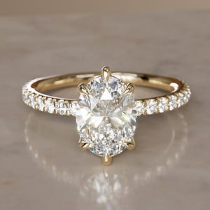 May include: A gold engagement ring featuring an oval-cut diamond centre stone. The band is embellished with smaller diamonds. The ring is displayed against a neutral backdrop, accentuating its brilliance and design. A classic symbol of love and commitment.