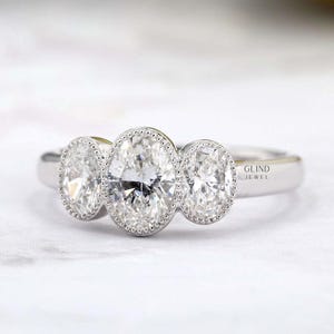 Peut inclure: Une bague de fiançailles à trois pierres avec des diamants de taille ovale. La bague présente un anneau en métal blanc et un sertissage en halo autour de chaque diamant. Les diamants scintillent sur un fond blanc. C'est un bijou.