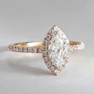 May include: A gold engagement ring featuring a marquise-cut diamond surrounded by a halo of smaller diamonds. The band is also set with small diamonds. The ring is displayed against a plain background. The text "GLIND JEWEL" is visible.