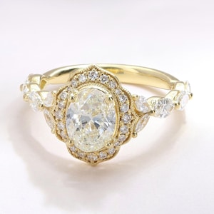 May include: A gold engagement ring featuring an oval-cut central diamond, encircled by a halo of smaller diamonds. The band incorporates further diamond embellishments. The ring is displayed against a white backdrop. The text "GLIND JEWEL" is visible.