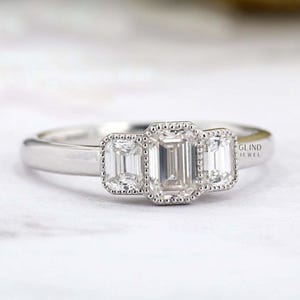 May include: A three-stone engagement ring with emerald-cut diamonds. The center diamond is flanked by two smaller stones, all set in a white metal band. The ring has a classic, elegant design.