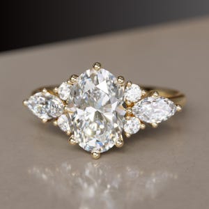 May include: A gold ring with an oval-cut central gemstone, flanked by smaller round and pear-shaped stones. The ring has a slender band and a cluster arrangement, creating a dazzling and refined appearance. The ring is a piece of jewellery.