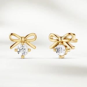 May include: A pair of gold-colored bow-shaped stud earrings. Each earring features a sparkling, round, clear gemstone at the center. The earrings are set against a soft, neutral background, highlighting their delicate design.