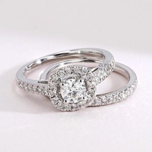 May include: Two silver engagement rings. The primary ring features a large, round, clear gemstone surrounded by smaller stones in a square halo. The band is also encrusted with small, clear stones. The second ring is a matching band with small, clear stones.