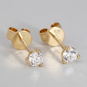 May include: A pair of gold stud earrings, each featuring a clear, round gemstone. The earrings have a post and back closure. The gemstones are held in place by a three-prong setting. The earrings are displayed against a light background. The text "GLIND JEWEL" is visible.