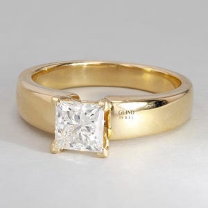 May include: A gold ring featuring a square-cut diamond. The band is a thick, polished gold. The diamond is held in place by four prongs. The ring is set against a white backdrop. The text "GLIND JEWEL" is visible on the side of the ring.