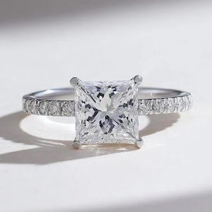May include: A silver engagement ring with a large, square-cut diamond as the centerpiece. The band is adorned with smaller, round diamonds. The ring is set against a white background, highlighting its sparkle and design.