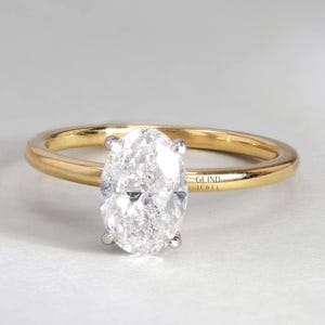 May include: A gold band engagement ring with a large, oval-cut diamond. The diamond is set with four prongs, and the band has a polished finish. The ring is set against a neutral background. The text "GLIND JEWEL" is visible.