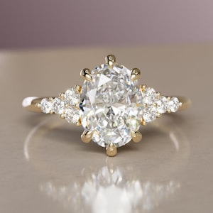 May include: A gold ring featuring a large, oval-cut diamond as the centerpiece, flanked by smaller, round diamonds. The ring has a delicate band and a cluster of accent stones on each side of the main stone. The ring is set against a neutral background.