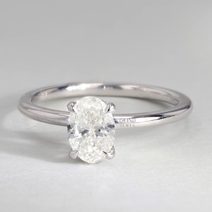 May include: A silver engagement ring with a large, oval-cut diamond as the centerpiece. The diamond is held in place by four prongs, and the band is thin and polished. The ring is set against a neutral background. The text "GLIND JEWEL" is visible.