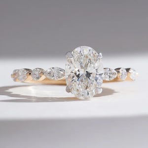 May include: A gold band ring with an oval-cut diamond center stone. The band features smaller, round and marquise-cut diamonds. The ring is set against a white background. The text "GLIND JEWEL" is visible on the ring.