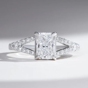 May include: A silver-coloured engagement ring with a radiant-cut diamond and a split shank. The ring has a large, rectangular centre stone and smaller diamonds set along the split band. The ring is set in a silver-coloured metal.