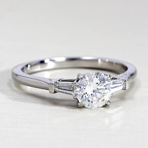 Peut inclure: Bague de fiançailles en argent avec un gros diamant rond et clair au centre. Deux diamants rectangulaires plus petits flanquent la pierre centrale. La bague a un anneau simple et poli.